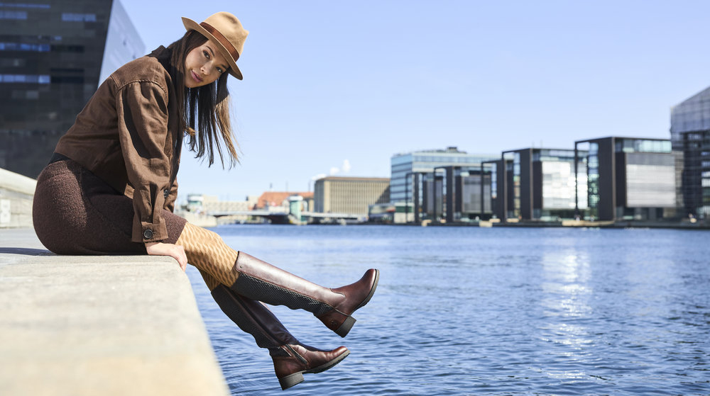 Kobieta siedzi na betonowym nabrzeżu nad wodą w miejskiej scenerii. Ma na sobie brązowy kapelusz, brązową kurtkę, legginsy i wysokie brązowe kozaki rieker. W tle nowoczesne budynki odbijają się w wodzie.